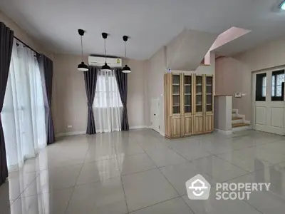 Spacious living room with gleaming tiled floors, elegant pendant lighting, and large windows draped in sheer curtains, complemented by a wooden bookshelf and staircase.