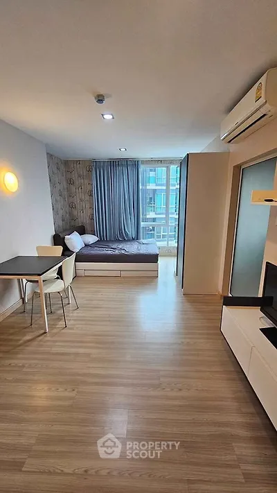 Modern studio apartment with wooden flooring, cozy bed, and dining area, featuring large windows for natural light.