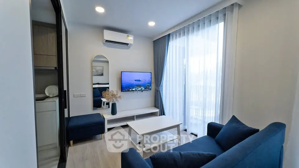Modern living room with blue sofa, wall-mounted TV, and large window with sheer curtains.
