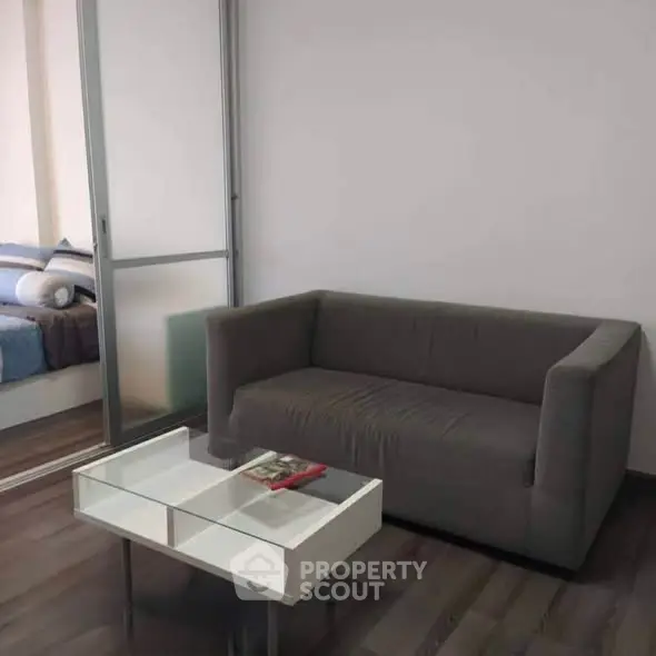 Modern living room with sleek gray sofa and glass coffee table, adjacent to a cozy bedroom.