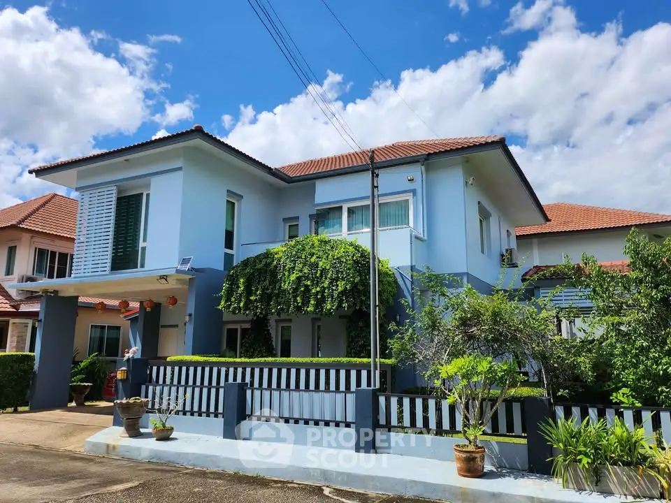 Charming blue two-story house with lush greenery and a fenced front yard under a bright blue sky.