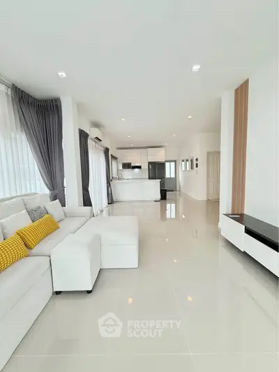 Spacious modern living room with open kitchen, featuring sleek white interiors and large windows for natural light.