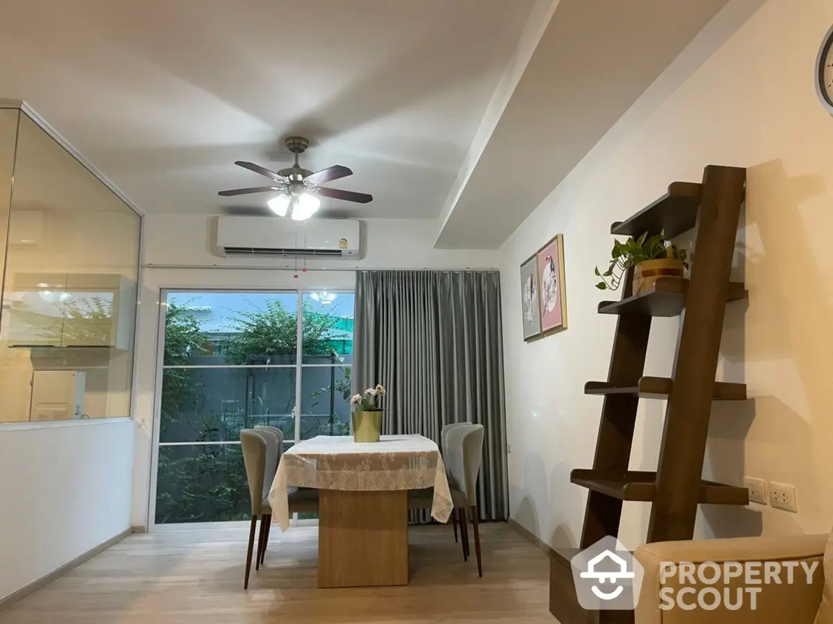 Spacious living room with modern ceiling fan, elegant dining set, and stylish wooden shelving leading to a cozy balcony with a serene view.