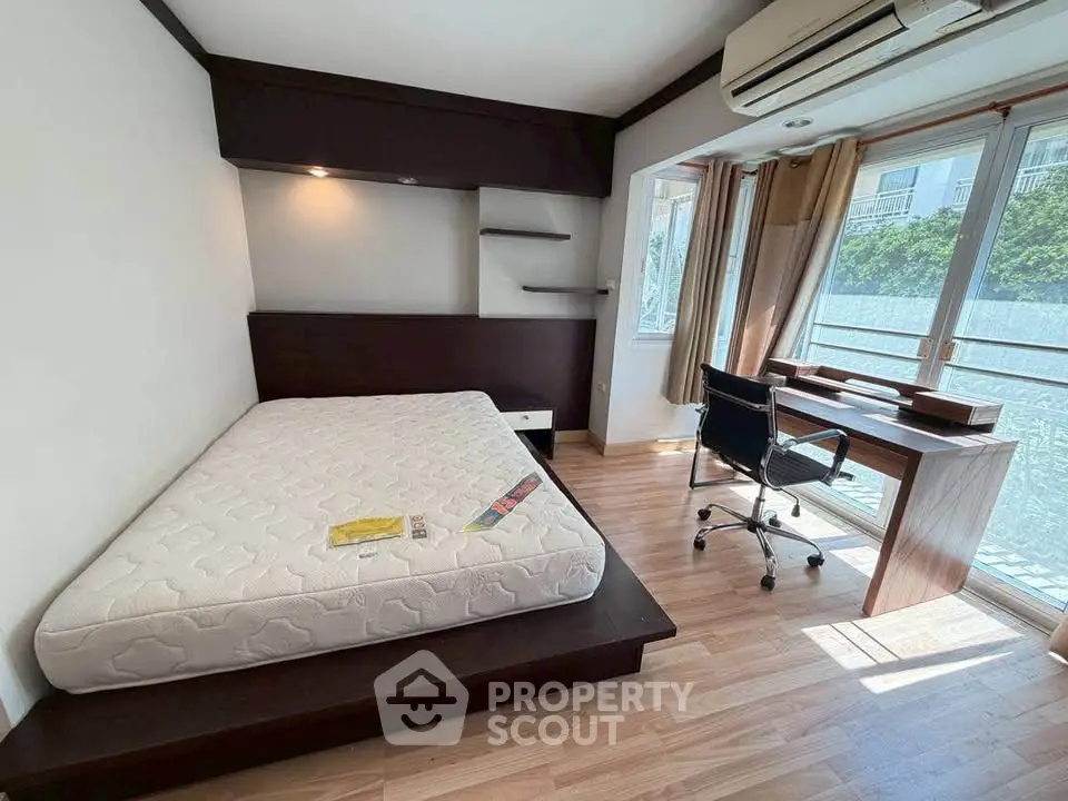 Modern bedroom with large window, wooden flooring, and workspace desk in a bright apartment.