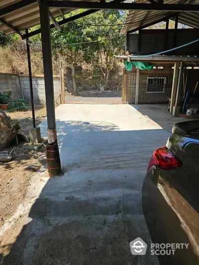 Spacious outdoor parking area with a protective metal roof, surrounded by natural greenery, offering ample space for multiple vehicles and outdoor activities.