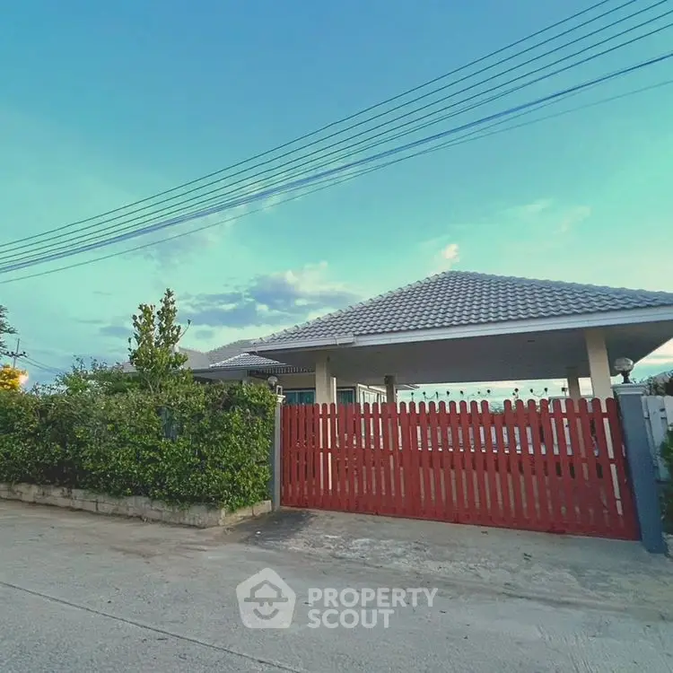 Charming suburban house with red gate and lush greenery under a clear blue sky.