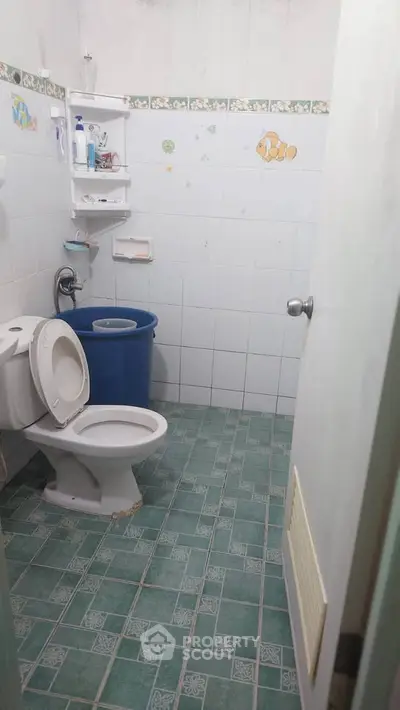 Cozy bathroom with green tiled floor and white fixtures, featuring a blue laundry bin and wall shelves.