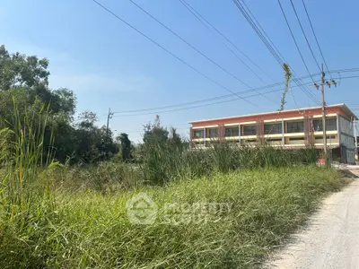 Spacious land plot with lush greenery and a nearby building under clear blue sky.