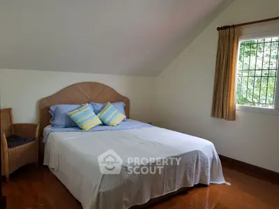 Cozy bedroom with sloped ceiling and natural light, featuring a comfortable bed and wicker chair.