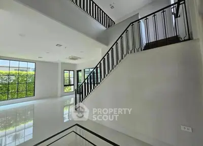 Spacious modern living room with elegant staircase and large windows offering lush green views.