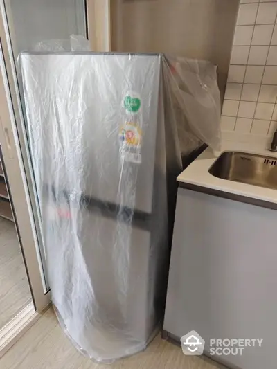 Modern kitchen with new fridge wrapped in protective plastic near sink and sliding door.