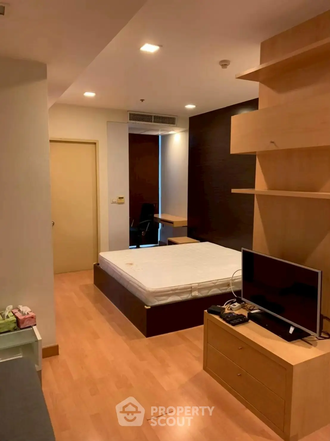 Modern studio apartment with wooden flooring and built-in shelves, featuring a cozy bed and flat-screen TV.