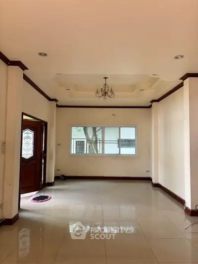 Spacious living room with elegant chandelier and polished tile flooring, featuring a wooden door and large window.
