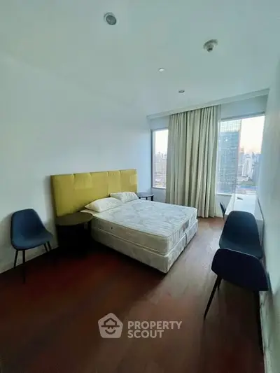 Modern bedroom with city view, featuring a cozy bed, stylish chairs, and large windows for natural light.