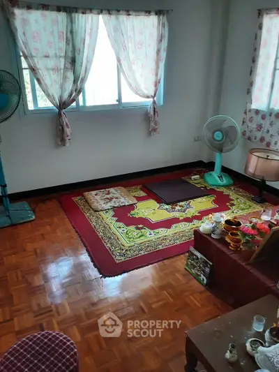 Cozy living room with wooden flooring and colorful rug, featuring a fan and natural light from windows.