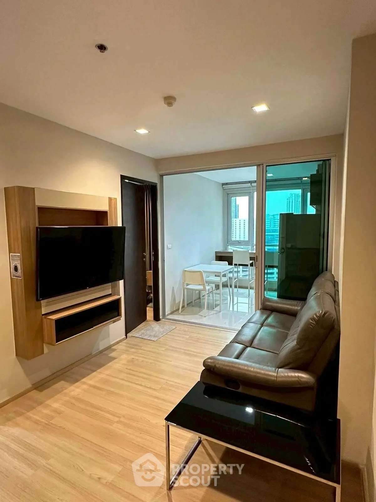Modern living room with sleek leather sofa and wall-mounted TV, leading to a bright dining area with city views.
