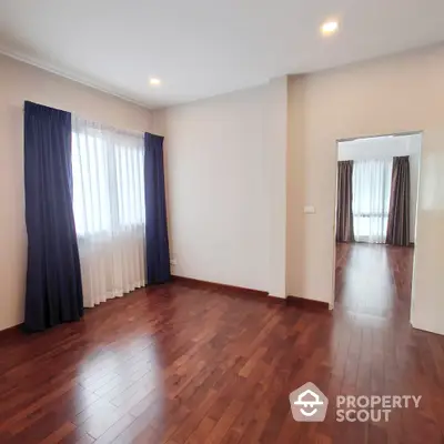 Spacious living room with polished hardwood floors, ample natural light, and elegant blue curtains, leading to an adjoining room through an open doorway.