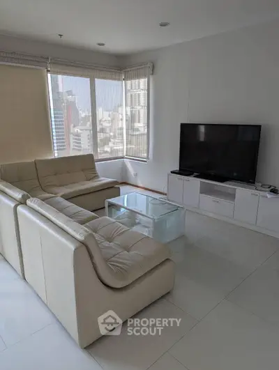Modern living room with large windows and city view, featuring a sleek white sofa and flat-screen TV.