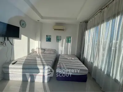 Bright bedroom with two beds, wall-mounted TV, and large windows with sheer curtains.