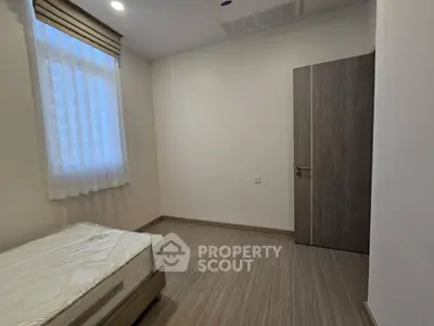 Minimalist bedroom with wooden flooring and natural light from window.