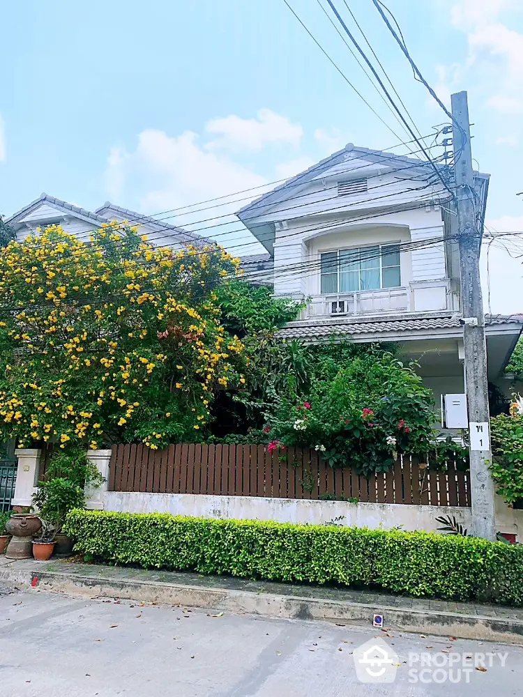 Charming two-story residence surrounded by lush greenery and vibrant flowering shrubs, featuring a classic design with a welcoming ambiance, nestled in a serene neighborhood.