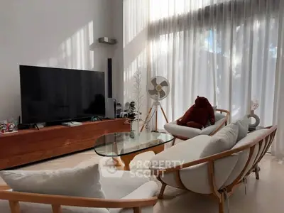 Stylish modern living room with large TV, elegant furniture, and natural light through sheer curtains.