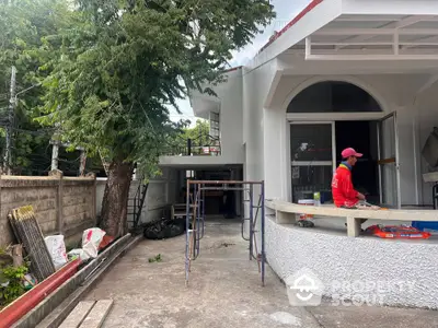 Charming white house exterior with spacious patio under renovation, surrounded by lush greenery.