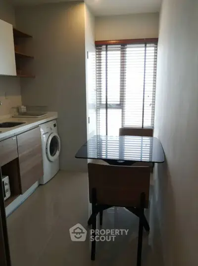 Cozy kitchen with washing machine and dining area, featuring natural light through large window blinds.