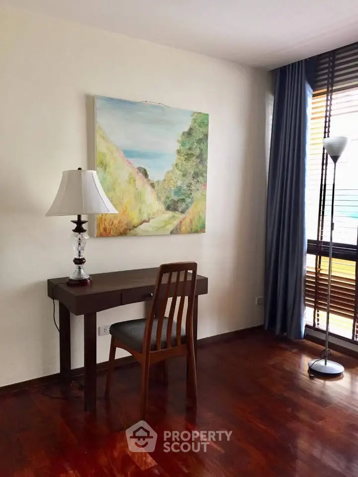 Charming study area with wooden desk, chair, and elegant lamp, featuring a large window with natural light.