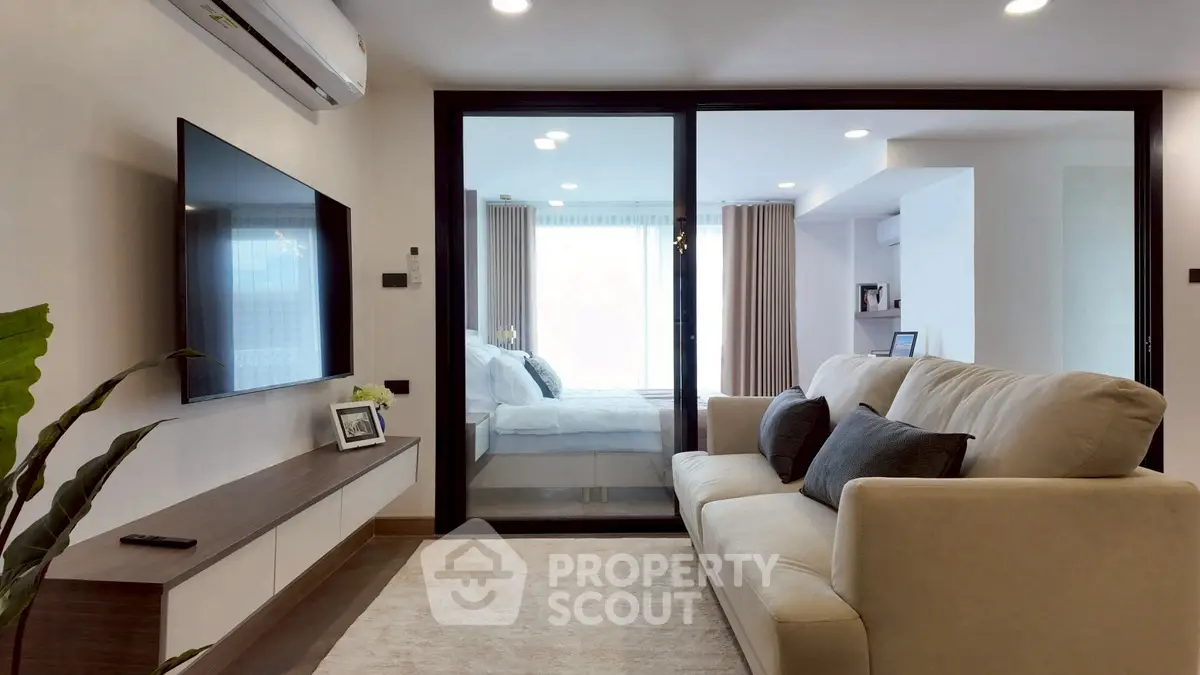 Modern living room with cozy sofa and large TV, leading to a bright bedroom through glass doors.