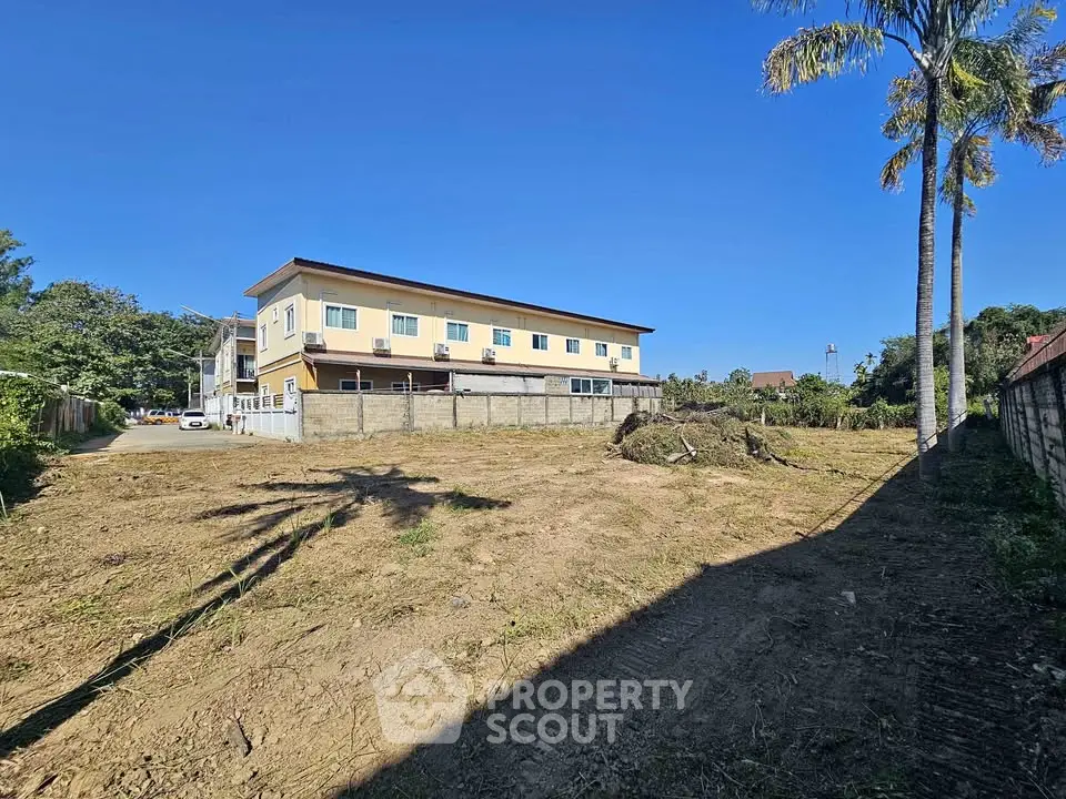 Spacious vacant land with potential for development, featuring a nearby building and lush greenery.