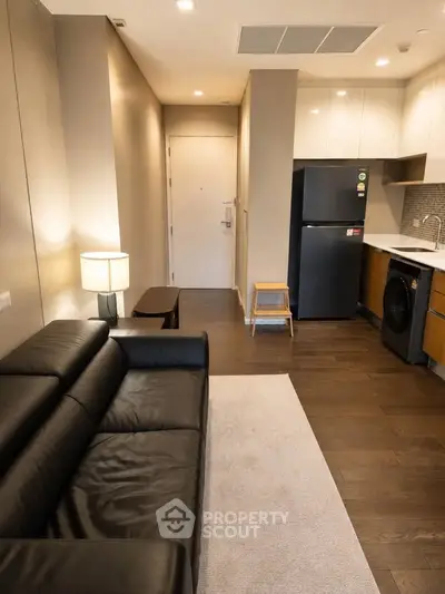 Modern apartment interior with sleek kitchen and cozy living area, featuring a black leather sofa and wooden flooring.