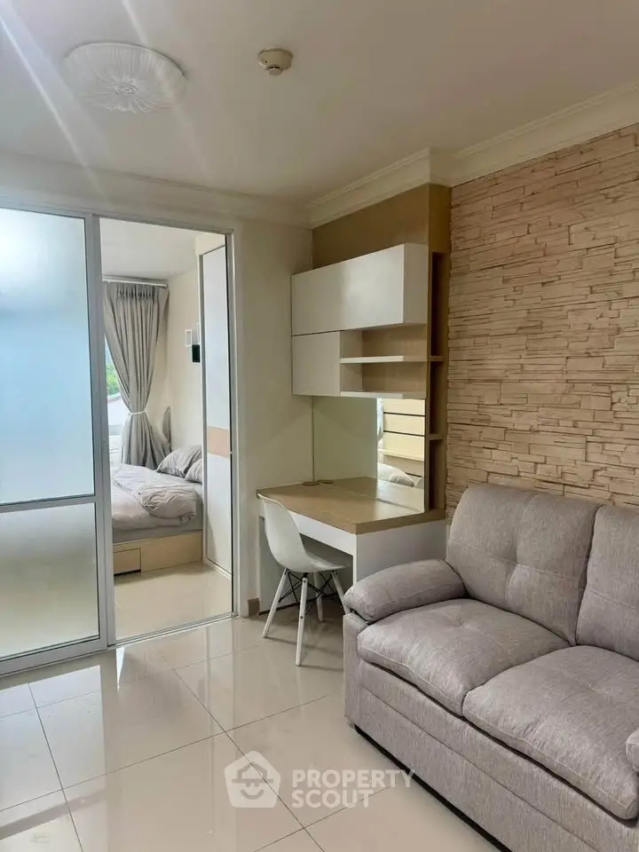Modern living room with cozy sofa and study area, adjacent to a bright bedroom with sliding glass doors.