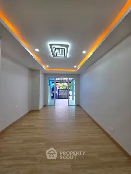 Spacious modern living room with stylish ceiling lights and wooden flooring, leading to a bright outdoor view.