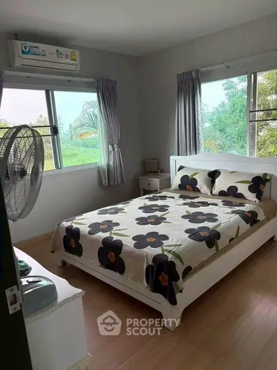 Cozy bedroom with floral bedding and large windows offering lush green views.