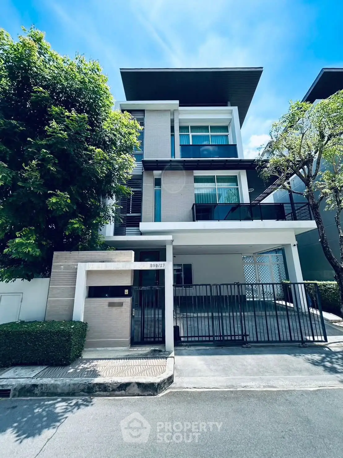 Modern three-story house with sleek design and gated entrance in a sunny neighborhood.