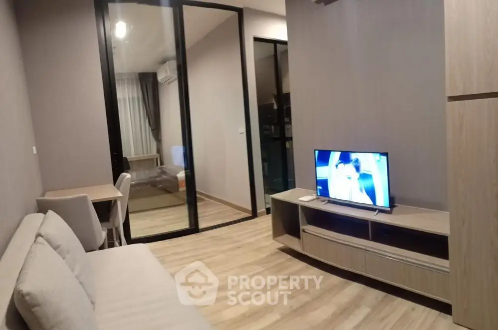 Modern living room with wooden flooring, cozy sofa, and TV, leading to a bedroom through glass doors.