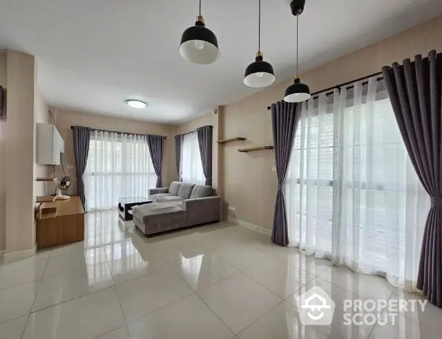 Spacious and elegantly designed living room with glossy tiled flooring, large windows draped with stylish curtains, and modern hanging light fixtures.