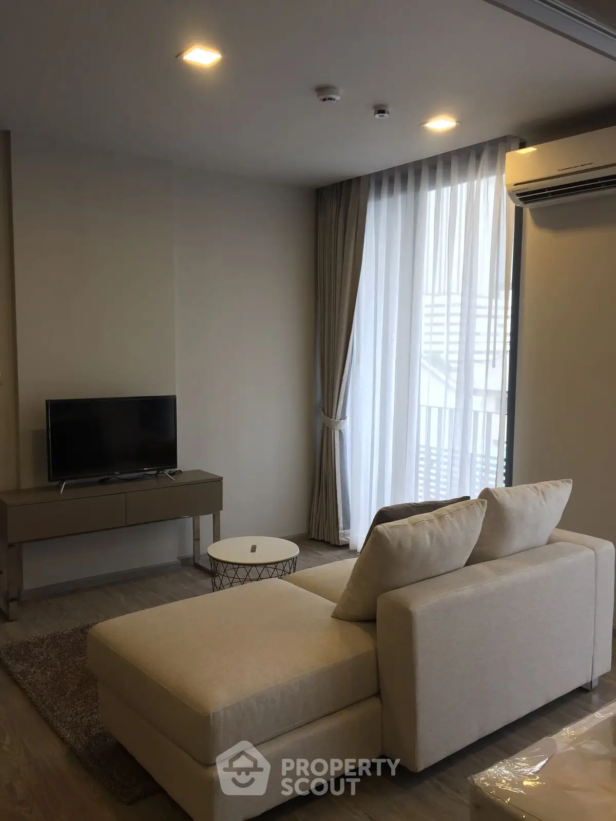 Modern living room with cozy beige sofa and flat-screen TV, featuring large window with sheer curtains.