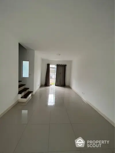 Spacious and bright living room with glossy tiled flooring, large windows draped with elegant curtains, and a modern staircase leading to an upper level.