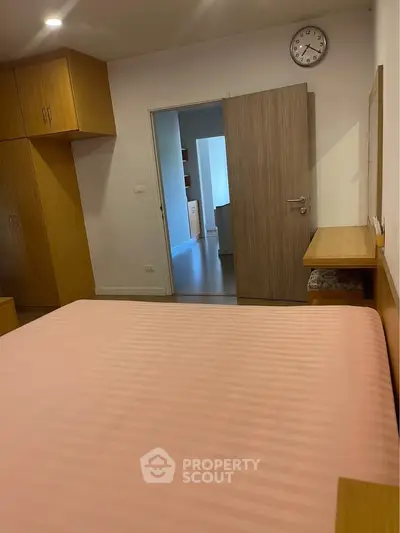 Cozy bedroom with wooden furniture and pink bedding, featuring a wall clock and open door to hallway.