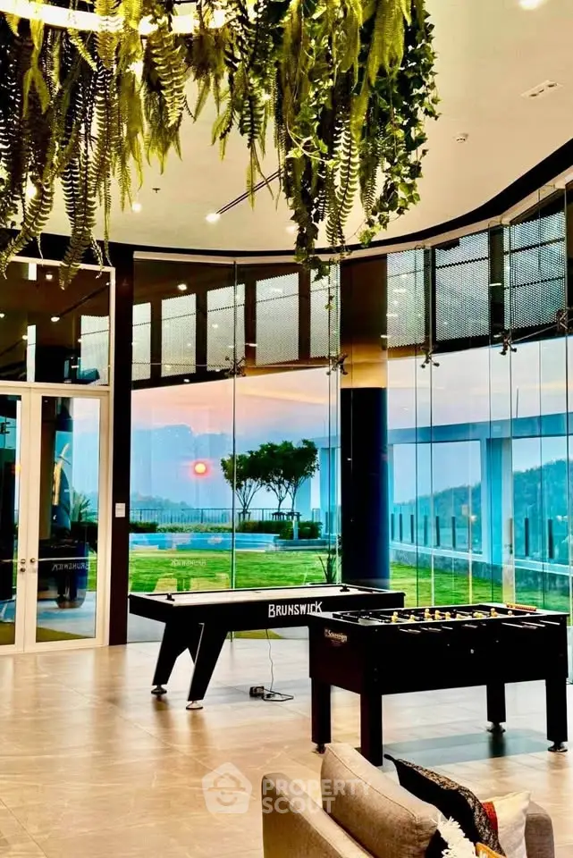 Luxurious modern interior with panoramic glass walls and game tables, offering stunning views of lush greenery.