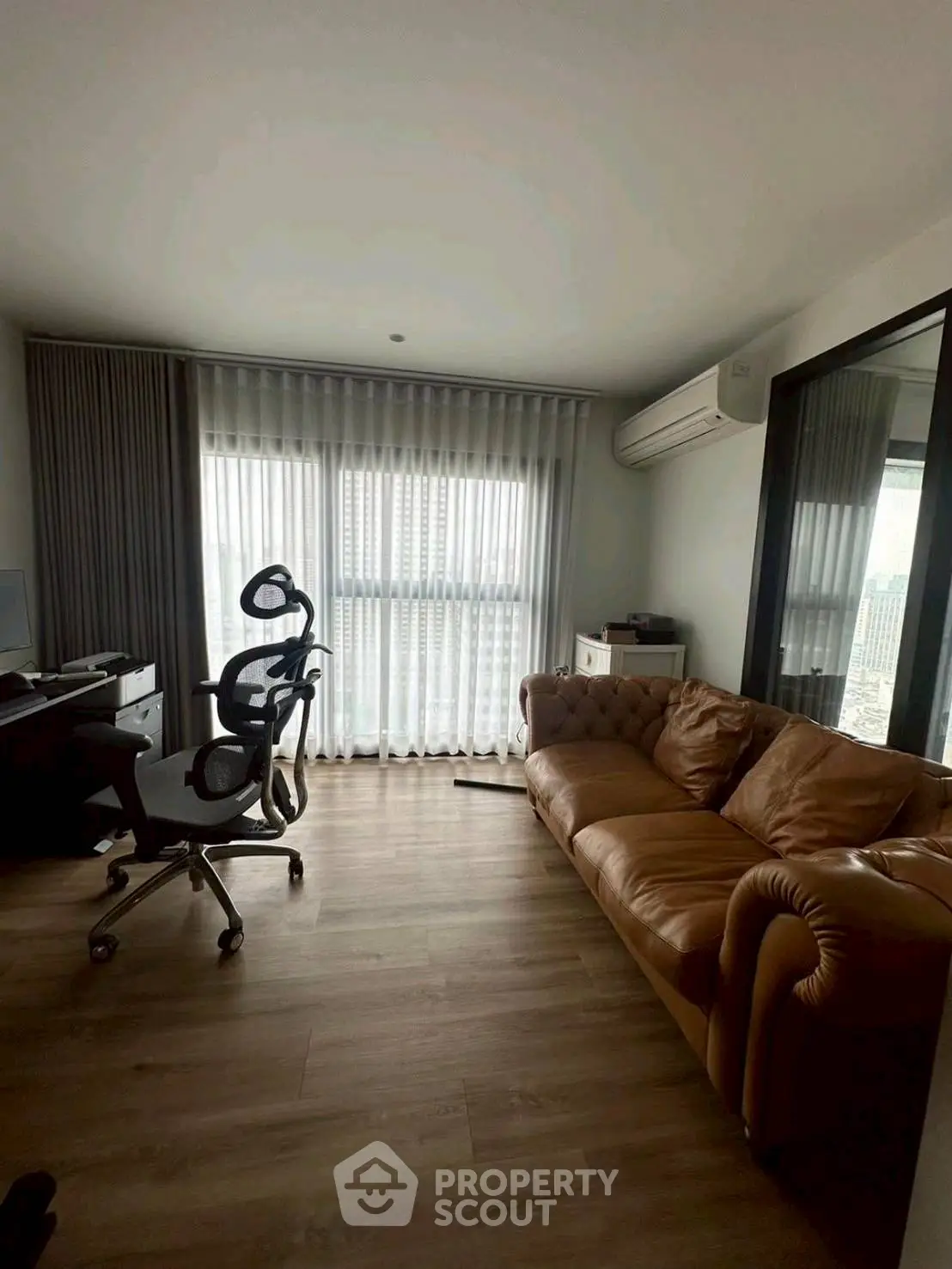 Modern living room with leather sofa and office setup, featuring large window and wooden flooring.