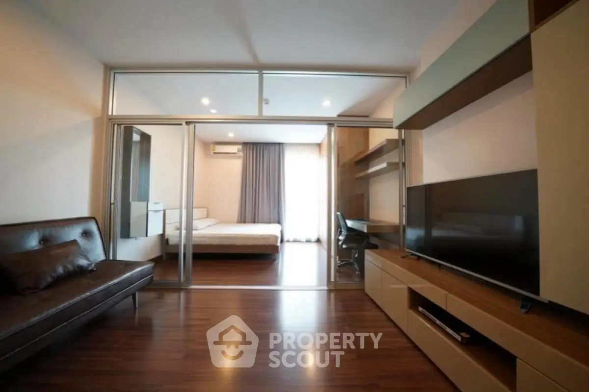 Modern living room with sleek furniture and large TV, leading to a cozy bedroom through glass doors.