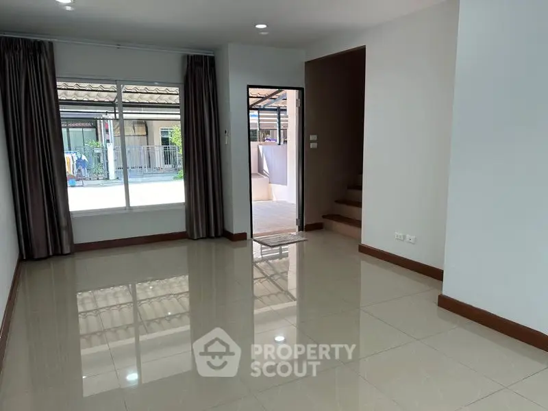Spacious and bright living room with glossy tiled flooring, large windows with curtains, and a view of the peaceful neighborhood.