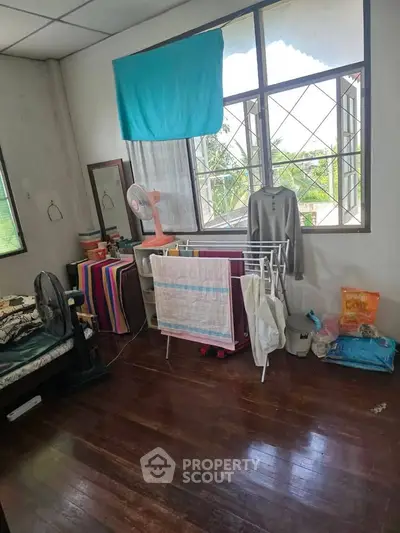Cozy bedroom with wooden floors, large windows, and natural light, featuring a drying rack and fan for comfort.
