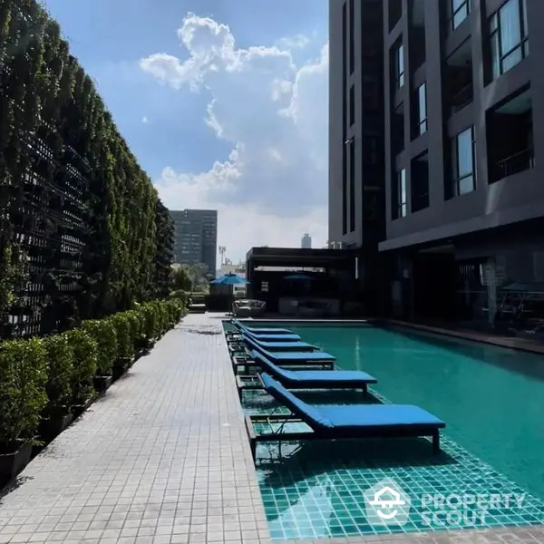 Luxurious urban oasis with a sparkling pool flanked by sun loungers and lush greenery, set against a modern residential building under a clear blue sky.