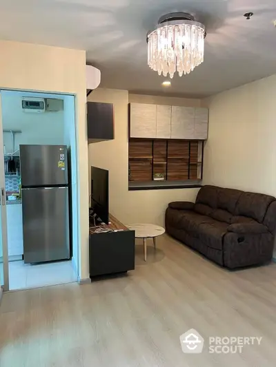 Elegant living room with modern furnishings, a plush sofa, and a sparkling chandelier, complemented by sleek wooden cabinetry and a full-sized refrigerator.