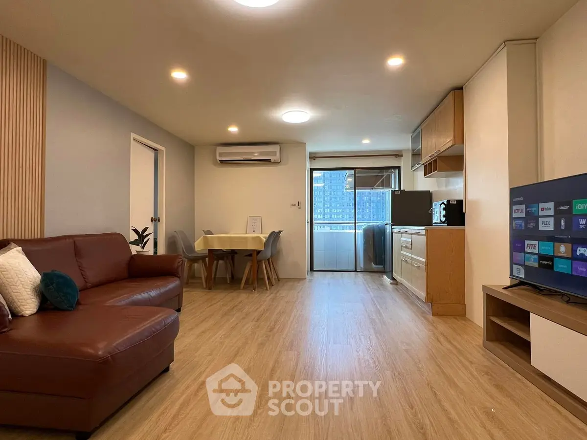 Modern living room with open kitchen and balcony view, featuring sleek furniture and wooden flooring.