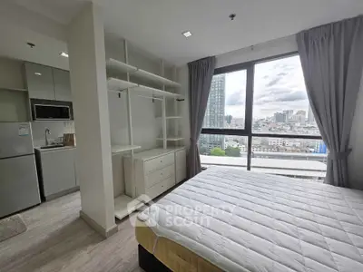 Modern bedroom with large window offering city view, adjacent to compact kitchen in urban apartment.